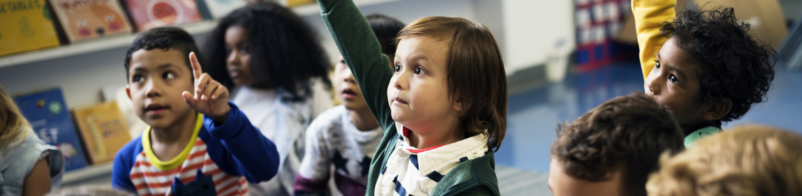 Young children in a classroom, one boy putting his hand up