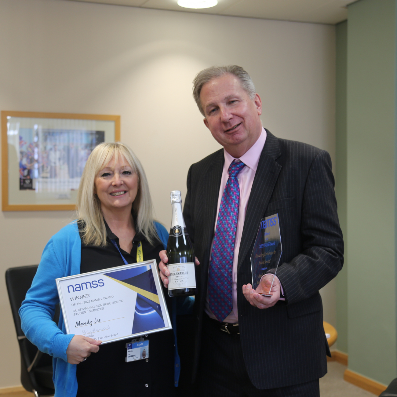 Mandy Lee holding an award, certificate and bottle of champagne stood next to Dr Paul Phillips CBE in his office