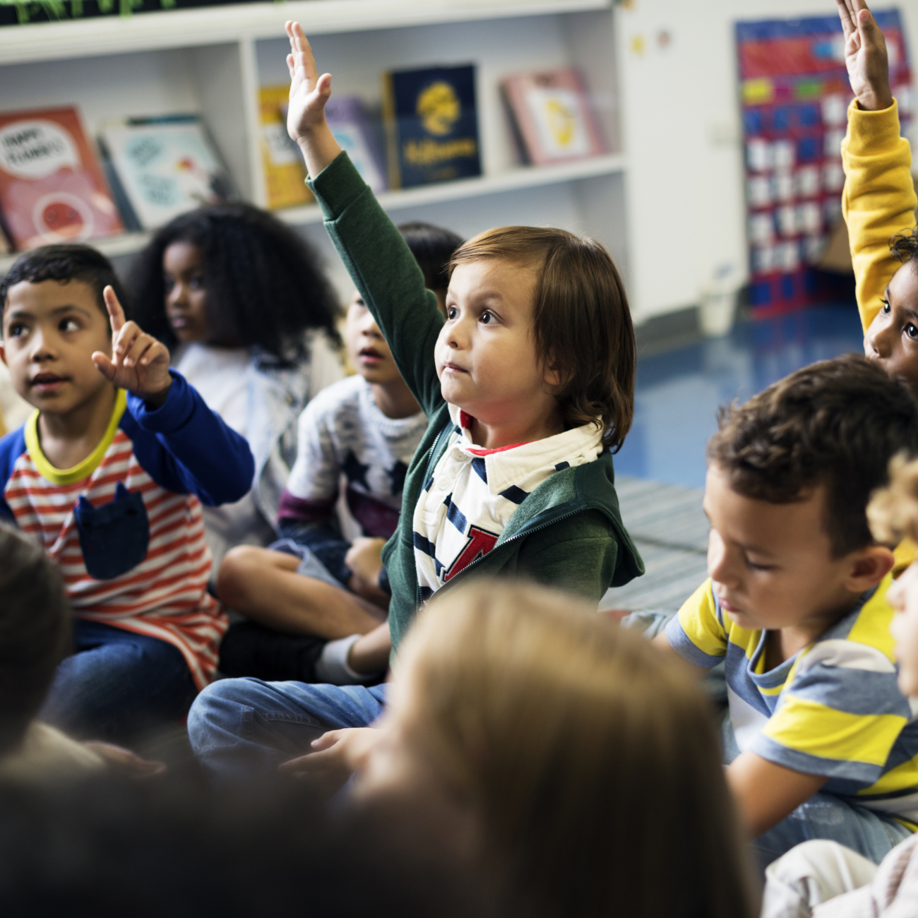 Young children in a classroom, one boy putting his hand up