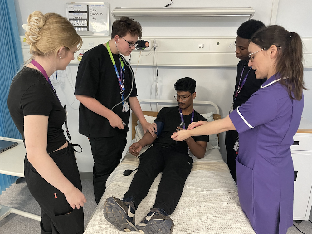 a students lies in a a hospital bed getting his blood pressure checked by other students